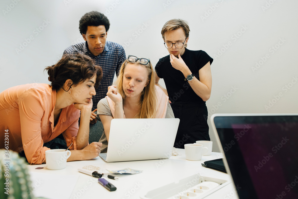 Multi-ethnic colleagues planning strategy while looking at laptop in illuminated office board room Multi-ethnic colleagues planning strategy while looking at laptop in illuminated office board room