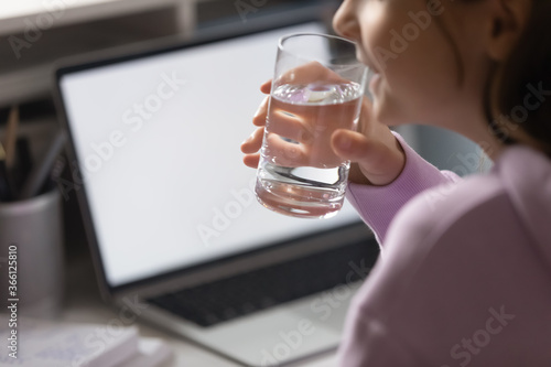 Close up thirsty teenage girl drinking pure mineral water, holding glass, teenager sitting at table with laptop with empty white screen mockup, healthy lifestyle and refreshment concept