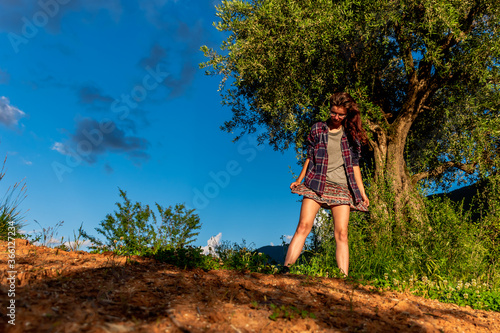 A full body shot of a happy young Caucasian redhead woman posing on a hiking path next to a tree in the mountains under warm summer sunlight  Puget-Theniers  France 