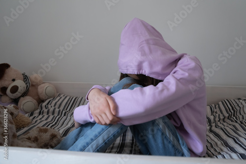 Lonely stressed teenage girl wearing hood sitting on bed alone, crying, thinking about troubles, hiding face, sad upset teenager feeling misunderstood and lonely, child and psychological problem