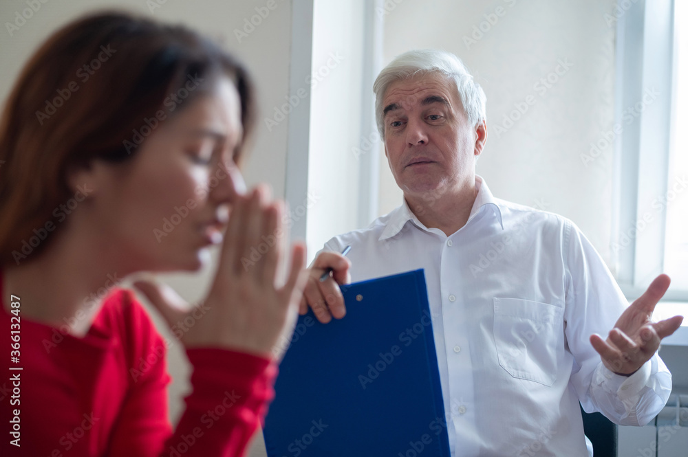 Obraz premium A frustrated crying woman holds palms in her face at a session with a male psychologist. Mature gray-haired psychotherapist talking to a female patient with depression and neurosis. Unbalanced psyche.