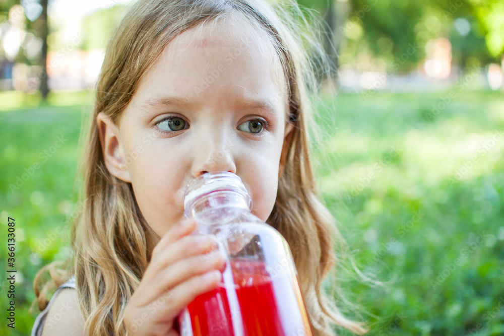 Portrait of a beautiful little girl with loose blond hair on a background of summer greenery. The child drinks juice with pleasure. Portrait of a beautiful little girl with loose blond hair on a background of summer greenery. The child drinks juice with pleasure.