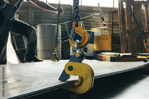 A worker in a factory lifts a bundle of sheet metal using a crane and mechanical clamps. Metal transportation at the plant. Clamping mechanism for sheet metal.