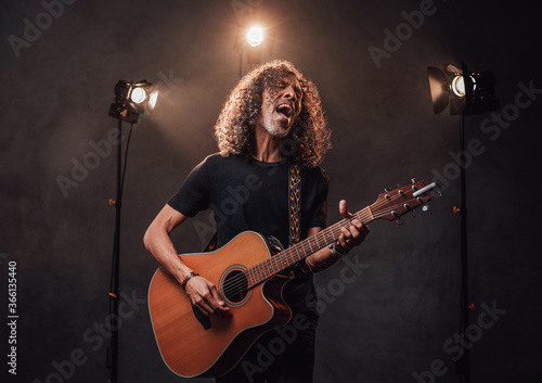 Middle aged hispanic musician in black t-shirt emotionally singing and playing guitar. View of musician in the spotlight