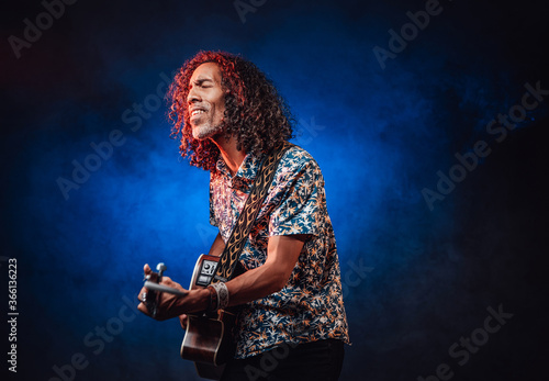 Middle aged hispanic man musician in a hawaiian shirt playing guitar on a dark illuminated by blue and red light. Concept of music, hobby, festival