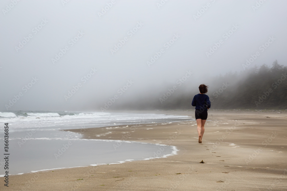 Athletic Caucasian Girl Running on a Sandy Beach near Pacific Ocean Coast during a cloudy and foggy morning. Taken at Raft Cove, Vancouver Island, British Columbia, Canada.