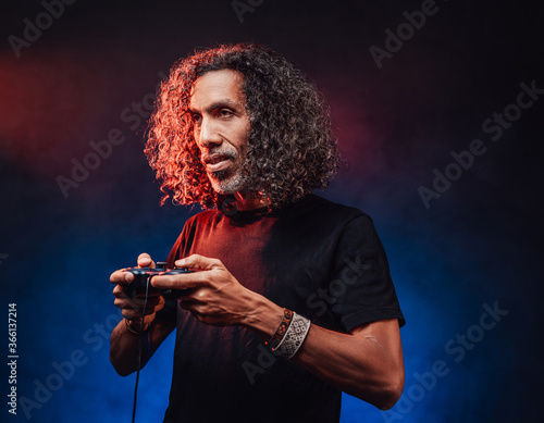 Middle aged hispanic male with long curly hair playing video game with joystick, looks focused. Hi-tech concept with vr glasses