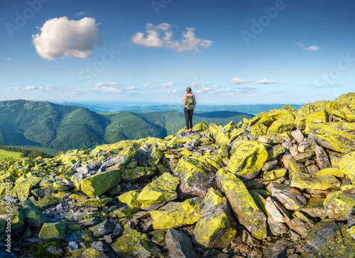Beautiful mountains and standing young woman with backpack on the green stones at sunset in summer. Landscape with sporty girl on the mountain peak, forest, hills , blue sky with clouds. Travel