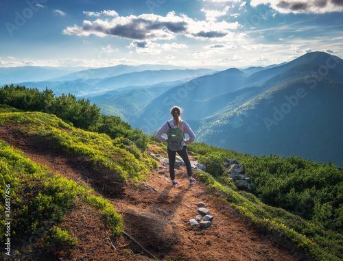 Beautiful mountains and standing young woman with backpack on the trail at sunset in summer. Landscape with sporty girl on the mountain peak, forest, hills , blue sky with clouds and sunbeams. Travel