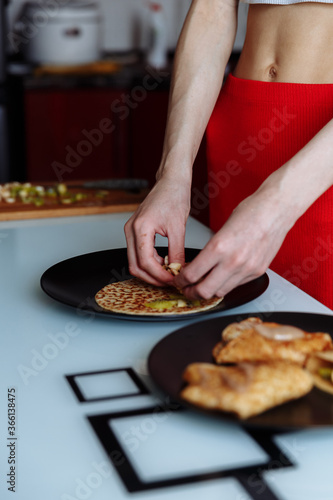 Woman's hand decorates pancakes with a fruits kiwi and banana. Black plate, white table