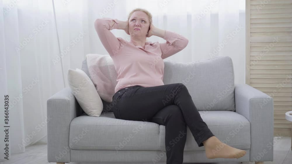 Unhappy senior woman sitting on couch with ears closed with hands. Wide
