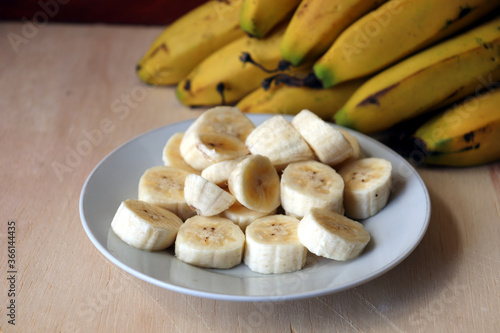 Banana slices on a white dish and bananas fruit in the background.