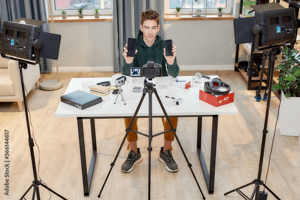 Full length shot of young male technology blogger recording video blog or vlog review of new smartphone, showing it and another one on camera at home studio. Blogging, Work from Home concept Full length shot of young male technology blogger recording video blog or vlog review of new smartphone, showing it and another one on camera at home studio. Blogging, Work from Home concept