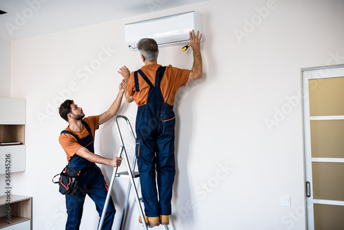 Two workers in uniform, air conditioning masters using ladder while installing a new air conditioner in the apartment. Construction, maintenance and repair concept photo