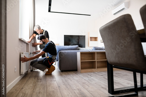 Картината върху платно Full length shot of two professional plumbers, workers in uniform fixing or installing heating radiator