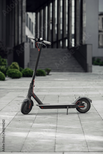 Nice black electrical scooter is parked near big glass building with offices.