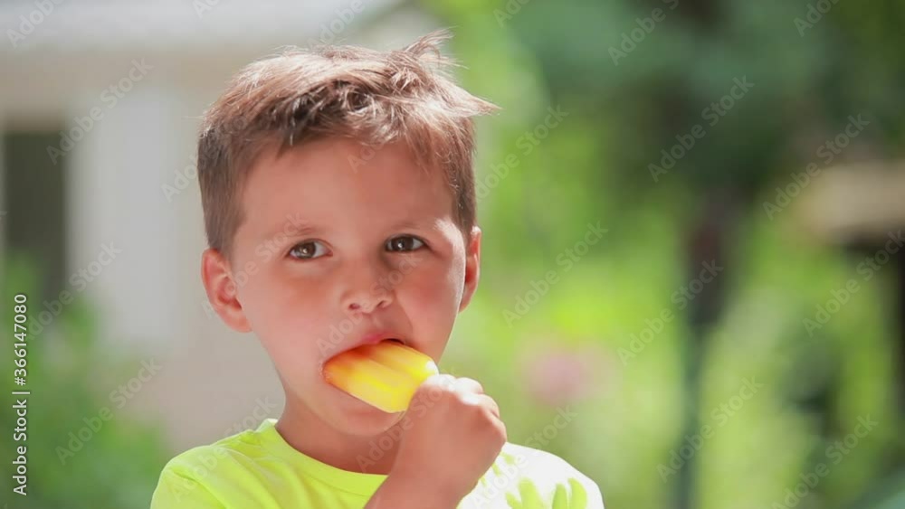 The cute cheerful boy is eating ice cream and showing big thumb outdoors on the sunny summer day The cute cheerful boy is eating ice cream and showing big thumb outdoors on the sunny summer day