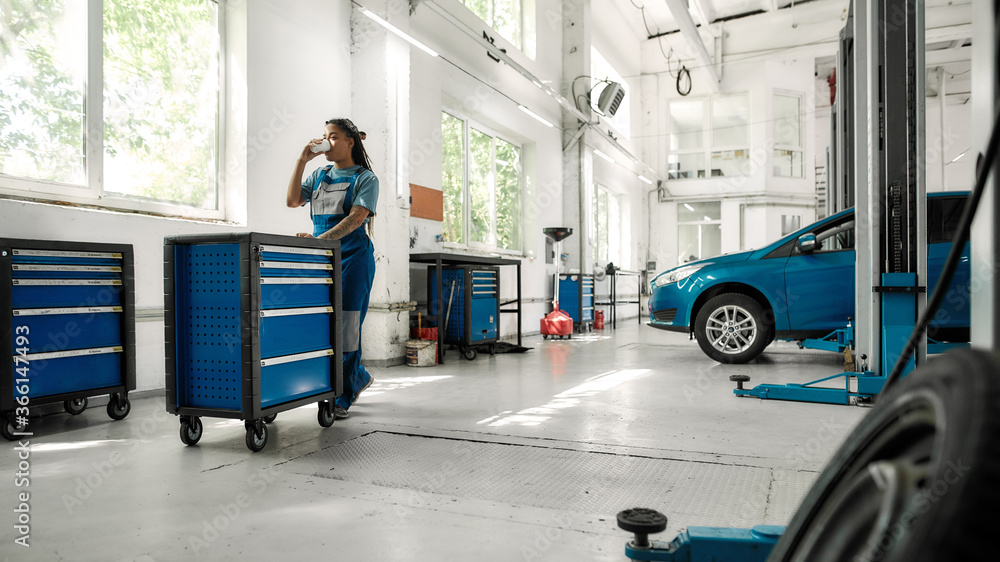 African american woman, professional female mechanic drinking coffee while pulling tool box cart in auto repair shop. Car service, maintenance and people concept African american woman, professional female mechanic drinking coffee while pulling tool box cart in auto repair shop. Car service, maintenance and people concept