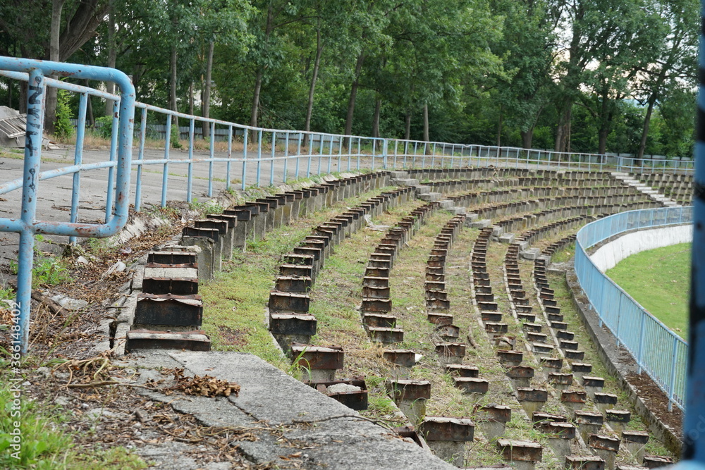 Obraz premium a football stadium tribune in disrepair or decayed in a provincial town in Eastern Europe. Wooden seats were removed because they were rotten and the space between rows is infested with weed.