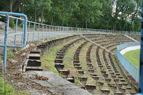 a football stadium tribune in disrepair or decayed in a provincial town in Eastern Europe. Wooden seats were removed because they were rotten and the space between rows is infested with weed. 