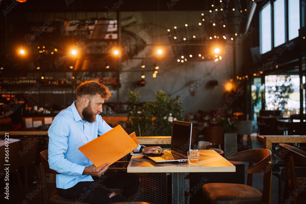 A young handsome caucasian businessman with a beard and a folder in his hand is sitting in a cafe and working on a laptop. Work in a restaurant A young handsome caucasian businessman with a beard and a folder in his hand is sitting in a cafe and working on a laptop. Work in a restaurant
