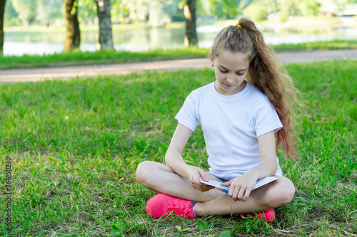 A school-age girl reading a book sitting on the grass in the park.