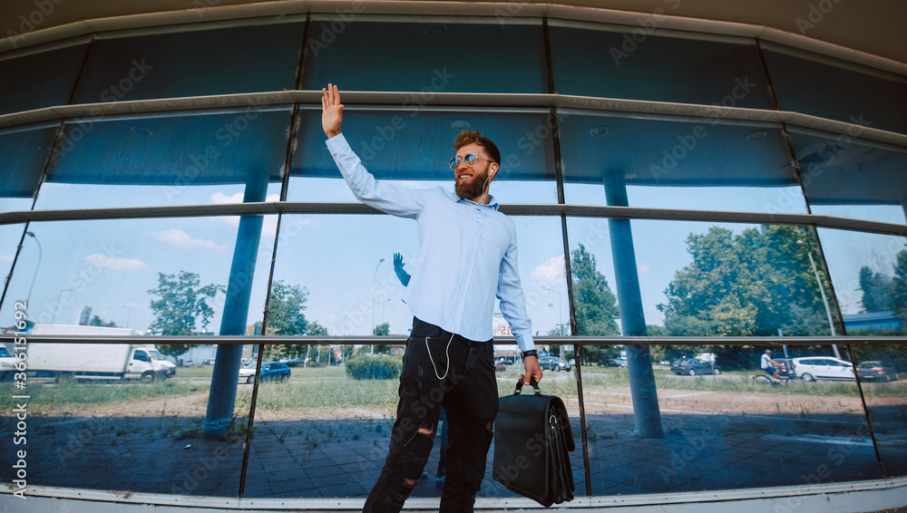 A young caucasian  businessman with sunglasses stands in front of the airport with a briefcase in his hand and waves