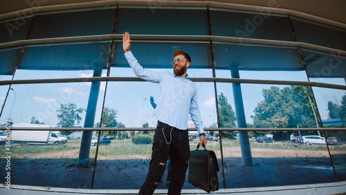 A young caucasian businessman with sunglasses stands in front of the airport with a briefcase in his hand and waves