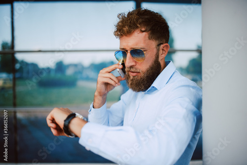 A young caucasian businessman with glasses stands in front of the company, looks at his watch and talks on the phone
