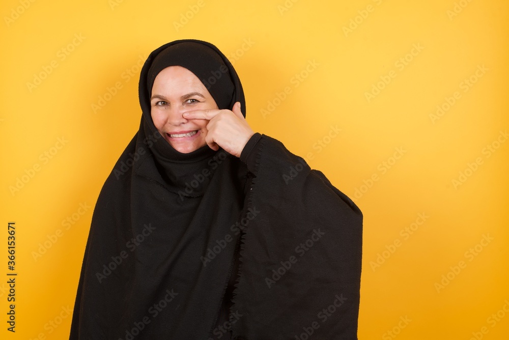 Beautiful middle aged muslim woman wearing black hijab over yellow background pointing unhappy to pimple on forehead, ugly infection of blackhead. Acne and skin problem Beautiful middle aged muslim woman wearing black hijab over yellow background pointing unhappy to pimple on forehead, ugly infection of blackhead. Acne and skin problem