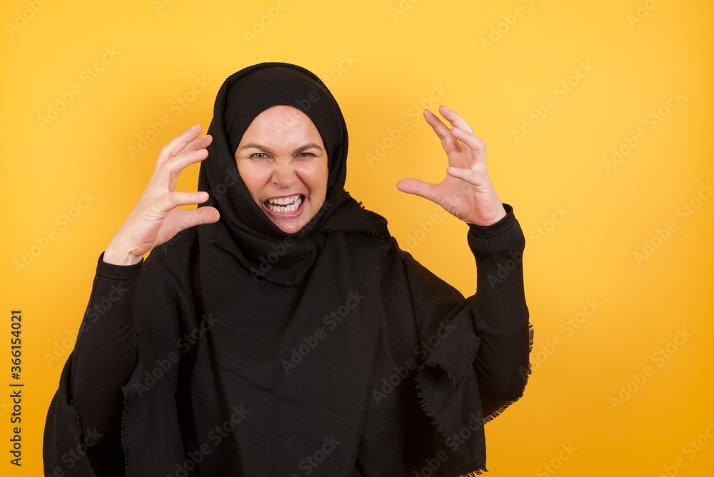 Beautiful middle aged muslim woman wearing black hijab over yellow background Shouting frustrated with rage, hands trying to strangle, yelling mad. Beautiful middle aged muslim woman wearing black hijab over yellow background Shouting frustrated with rage, hands trying to strangle, yelling mad.