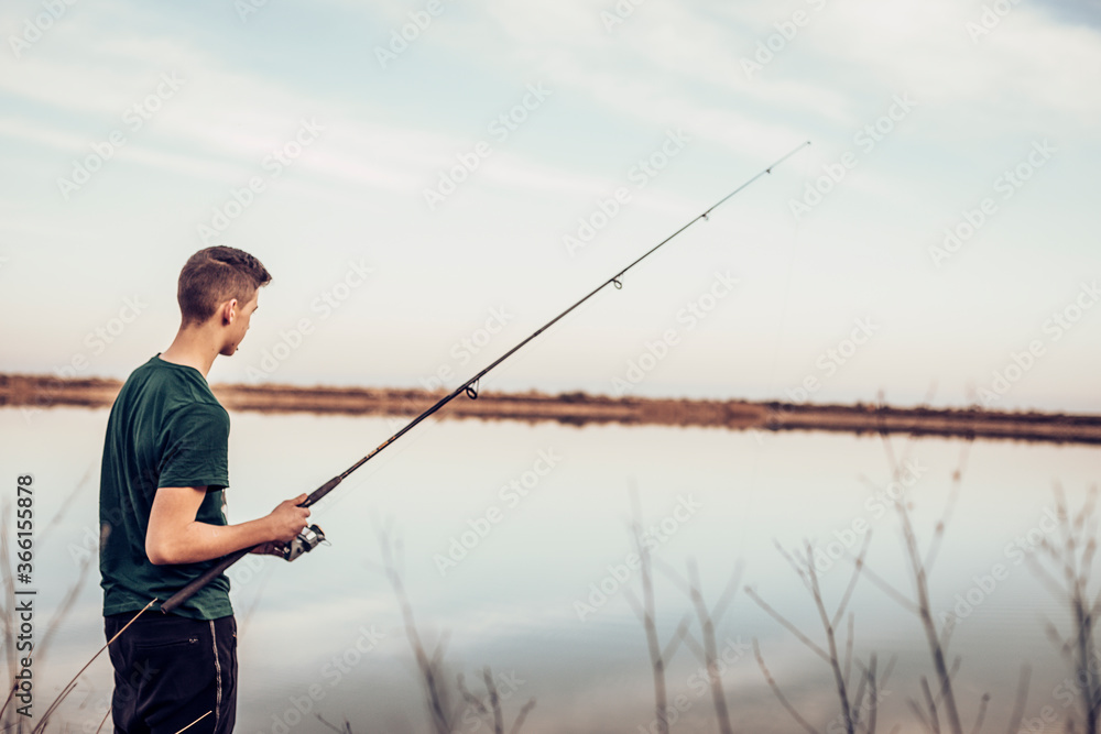 Side view of teenage boy fishing with rod in lake while standing against cloudy sky Side view of teenage boy fishing with rod in lake while standing against cloudy sky