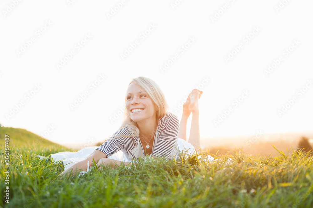 Happy thoughtful young woman lying on grassy land against clear sky in park during sunset Happy thoughtful young woman lying on grassy land against clear sky in park during sunset