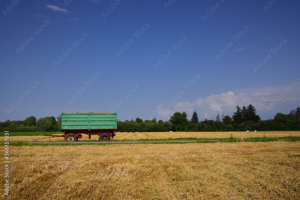 In summer, next to a harvested grain field, there is a green agricultural trailer against a blue sky