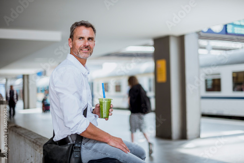 Thoughtful businessman holding drink looking away while sitting at railroad station platform