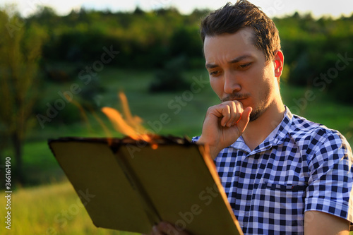 White Caucasian young man or adult guy reading a burning book on blurred forest background. Serious thoughtful face photo