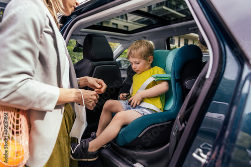 Little boy fastening seat bell of child's seat in car while his mother watching him