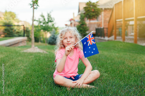 Adorable cute happy Caucasian girl holding Australian flag. Smiling child sitting on grass in park holding Australia flag. Kid citizen celebrating Australia Day holiday in January outdoors.
