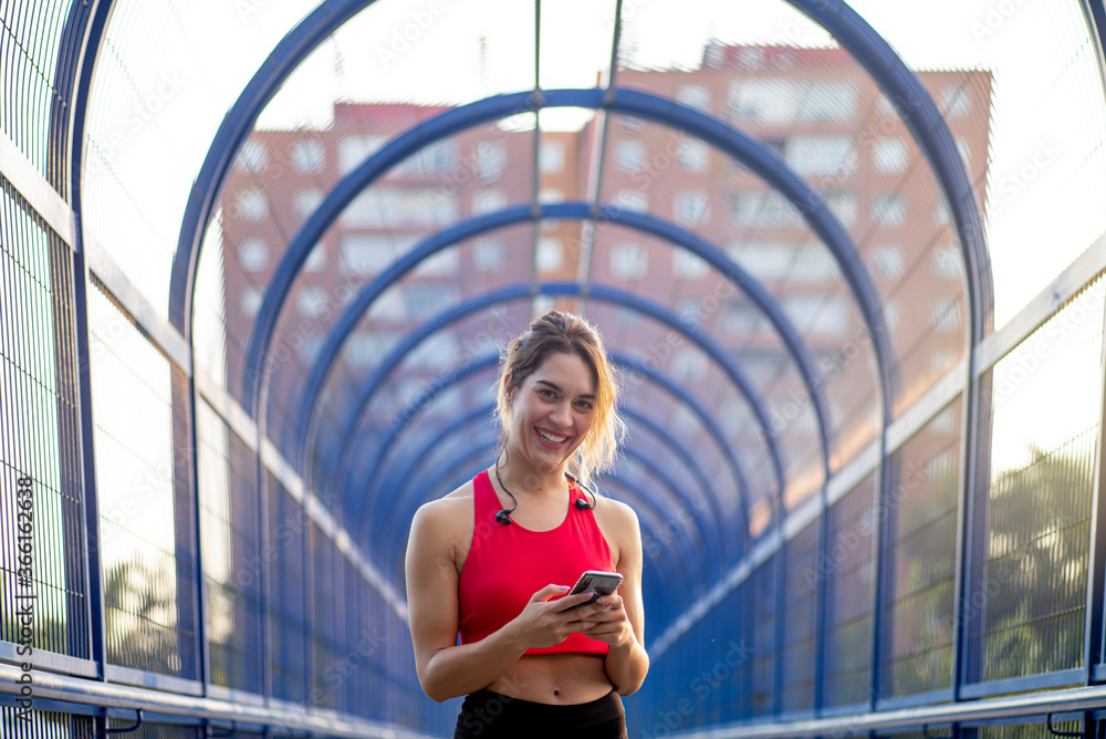 Portrait of smiling confident female athlete using smart phone while standing on covered bridge in city Portrait of smiling confident female athlete using smart phone while standing on covered bridge in city