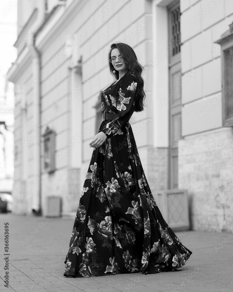 Young beautiful lady wearing black dress with colorful floral design and standing at city street. Pretty woman outdoor full length portrait. Fashion model.
