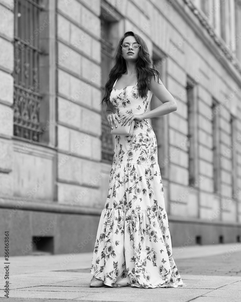 Young beautiful woman with long brunette wavy hair walking at city street. Elegant female wearing long colorful white and red floral dress. Girl in ball gown. Full length fashion portrait Young beautiful woman with long brunette wavy hair walking at city street. Elegant female wearing long colorful white and red floral dress. Girl in ball gown. Full length fashion portrait