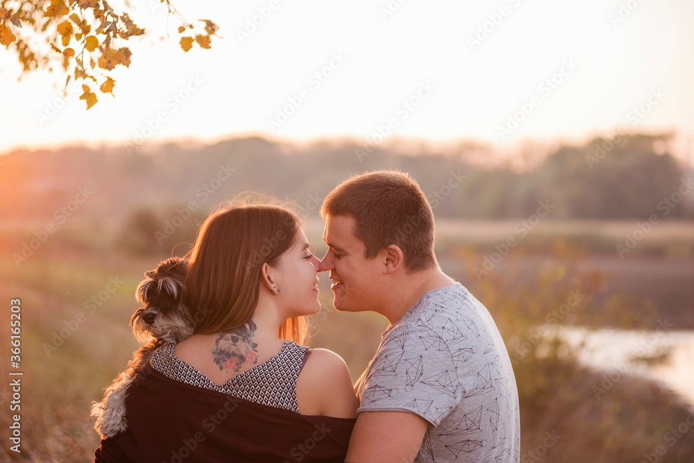A couple in love hugs, kisses in the rays of the autumn sun, with their backs turned to the camera, holding a Schnauzer in their arms. A beautiful girl has a dog portrait tattoo on her back. Family