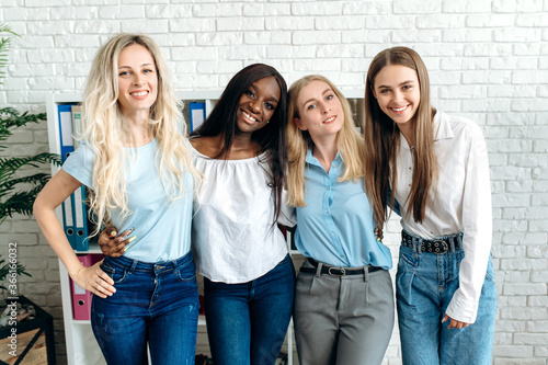 Attractive young smiling girls. Positive stylish female employees of different ethnicities are standing in their office in a cheerful mood looking at the camera and smiling