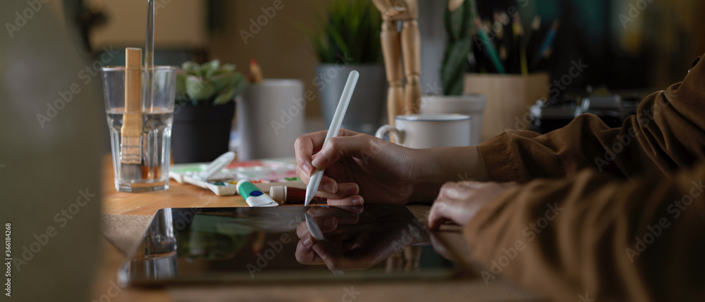 Female designer working on digital tablet while sitting at workspace with painting tools and supplies