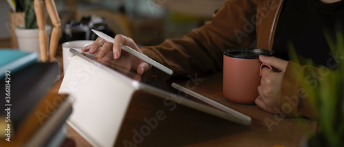 Female student using tablet while holding coffee cup on worktable with stack of books and supplies