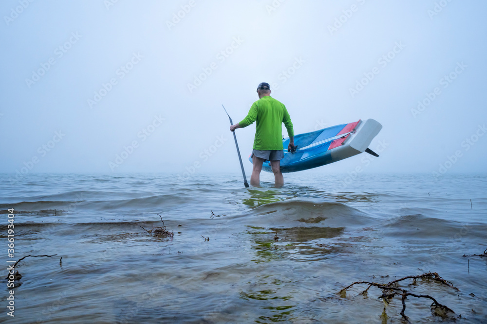 Naklejka premium mature male paddler is launching stand up paddleboard on a foggy lake - Boyd Lake State Park in northern Colorado, a popular boating and recreation destination.