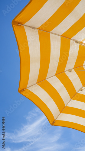 White and yellow striped beach umbrella. Blue sky in the background. View from below. Relaxing context. Summer holidays by the sea. General contest and location