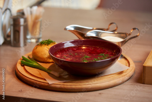 A photo below of a Ukrainian borsch with a pampushka, onion, and sour cream on a wooden tray on the table in a restaurant.