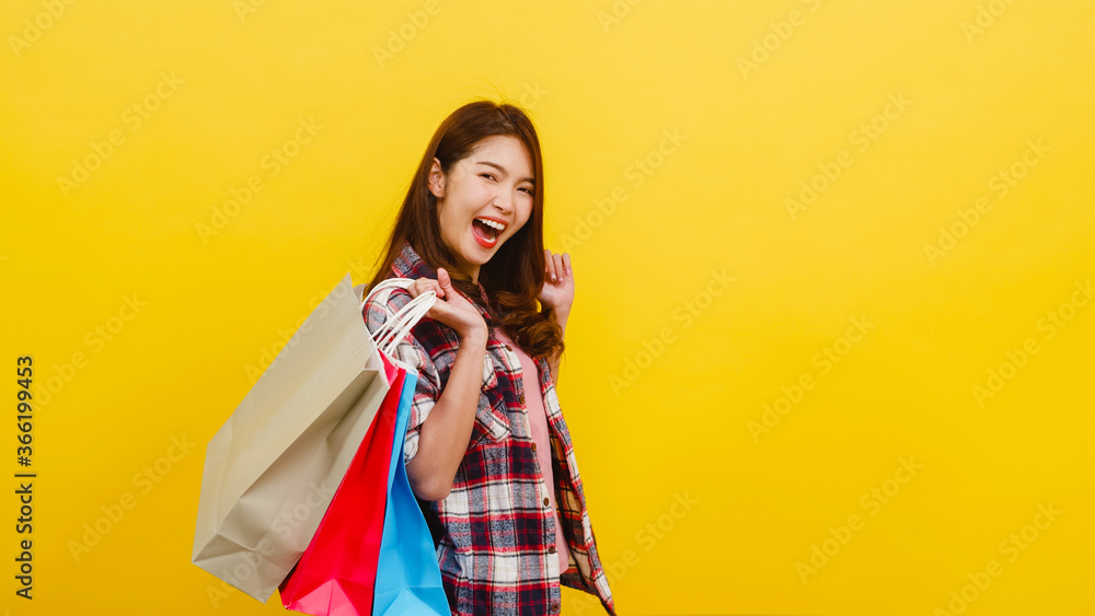 Happy excited young Asian lady carrying shopping bags with hand raising up in casual clothing and looking at camera over yellow background. Facial expression, seasonal sale and consumerism concept. Happy excited young Asian lady carrying shopping bags with hand raising up in casual clothing and looking at camera over yellow background. Facial expression, seasonal sale and consumerism concept.