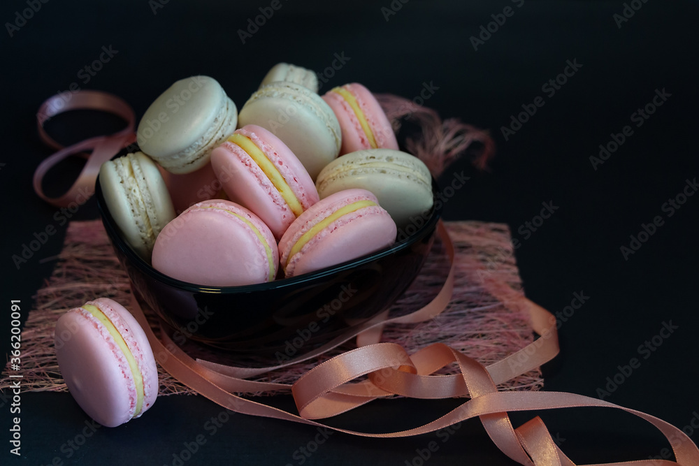French pink and cream colors macarons on black plate decorated by ribbon on dark background. French pink and cream colors macarons on black plate decorated by ribbon on dark background.